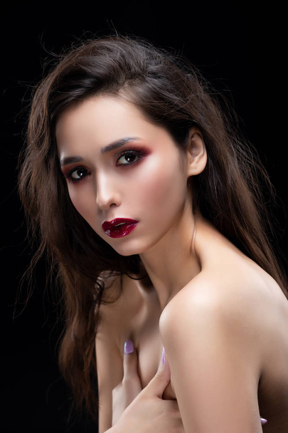 Portrait of a beautiful glamorous brunette girl sensually covers her naked breast with her hands. Shimmering makeup, red lips. Healthy smooth skin. Isolated on black. Copy space. - Фото, изображение