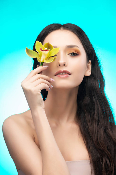 A beautiful brunette nude shoulders girl sensually holds a flower of an orchid in her hands. Nude make-up. Healthy smooth skin. Sky blue background. Copy space. Commercial and advertising design. - Foto, Imagen