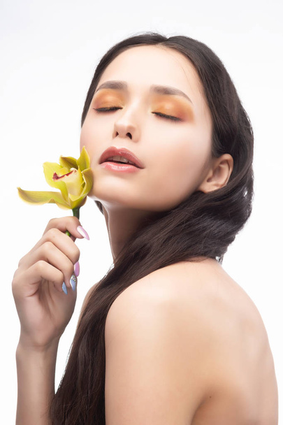 A beautiful brunette nude shoulders girl sensually holds a flower of an orchid in her hands. Nude make-up. Healthy smooth skin. Isolated on white background. Copy space. Commercial and advertising. - Photo, image