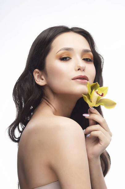 A beautiful brunette nude shoulders girl sensually holds a flower of an orchid in her hands. Nude make-up. Healthy smooth skin. Isolated on white background. Copy space. Commercial and advertising. - Photo, Image