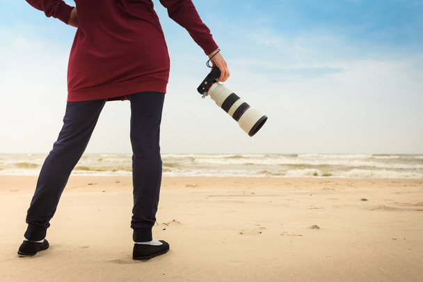 Unrecognizable woman having leisure time walking on beach during autumnal weather holding professional photography camera. - Photo, Image