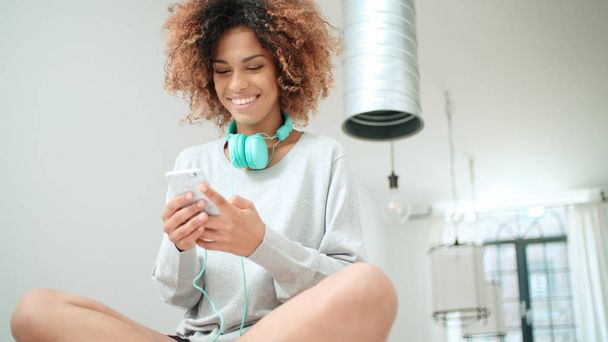 Happy and cheerful young woman enjoying music and holding phone. Beautiful young afro american woman with colorful headphones sitting on a kitchen table at home. - Photo, Image
