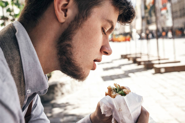 κομψό hipster άνθρωπος δαγκώνει σάντουιτς κάθεται σε ηλιόλουστο δρόμο. γρήγορη έννοια των τροφίμων. χώρο για το κείμενο - Φωτογραφία, εικόνα
