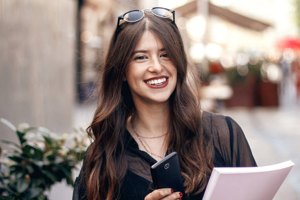 beautiful stylish hipster girl texting on phone, smiling in sunny city street, holding magazine.  - Photo, Image