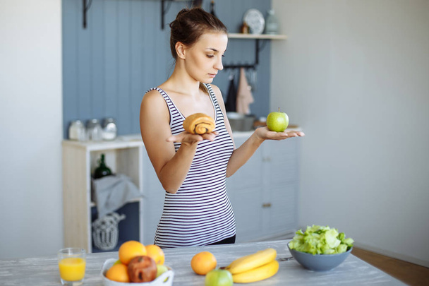 Woman holds in hand cake sweet bun and apple fruit choosing, trying to resist temptation, make the right dietary choice. - Foto, Bild
