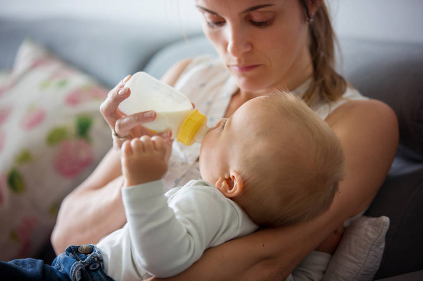 Mother, feeding her baby boy from bottle, comfortably sitting on the couch at home - Photo, image