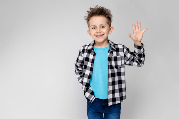 A handsome boy in a plaid shirt, blue shirt and jeans stands on a gray background. The boy smiles and shows the palm - Photo, Image
