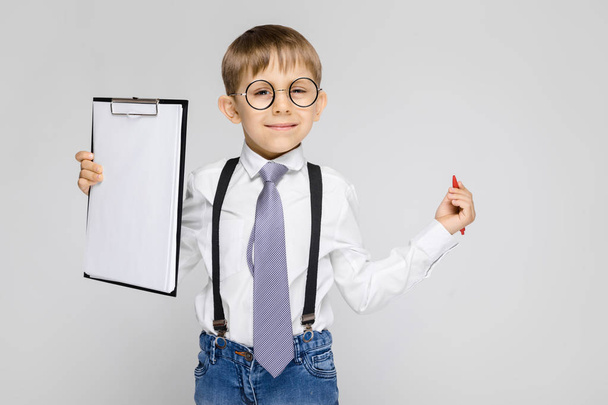 Ein charmanter Junge in weißem Hemd, Hosenträgern, Krawatte und heller Jeans steht vor grauem Hintergrund. der Junge hält einen Stift und Blätter für Notizen - Foto, Bild