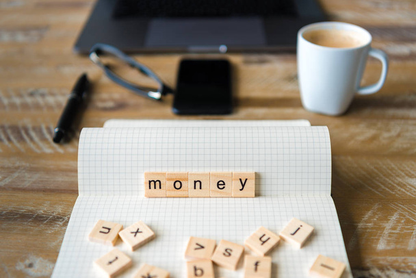 Closeup on notebook over wood table background, focus on wooden blocks with letters making Money word. Business concept image. Laptop, glasses, pen and mobile phone in a defocused background. - 写真・画像