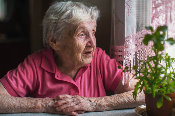 窓の外見て悲しいことにテーブルに座っている年配の女性. - 写真・画像
