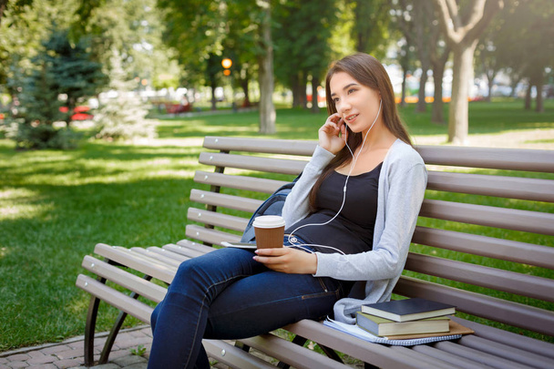 Thoughtful young female IT student in headphones. Listening to music and drinking coffee in the park - Foto, imagen