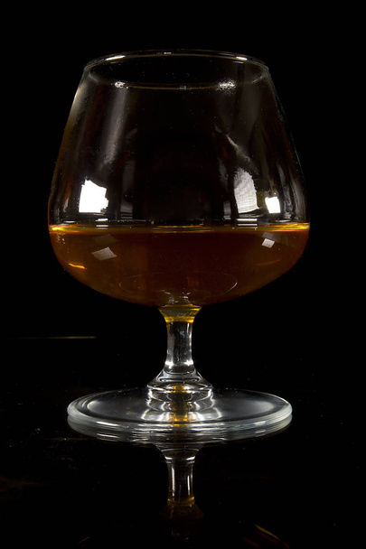 A glass of cognac on a black background on a reflective surface - Foto, afbeelding