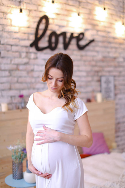 Pregnant american woman wearing white dress holding belly, inscription love on brick wall. - Photo, Image