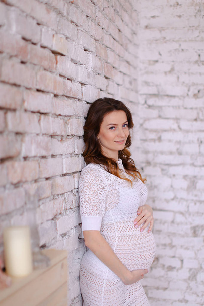 Mulher branca grávida vestindo vestido branco em pé no fundo da parede de tijolo e segurando barriga
. - Foto, Imagem