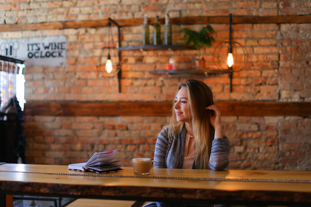 Χαλαρή κορίτσι που κάθεται κοντά σε πίνακα με φλιτζάνι καφέ και σημειωματάριο στο φιλόξενο café. - Φωτογραφία, εικόνα