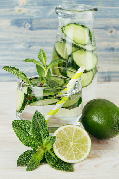 Cool transparent summer lemonade in glass with straw, bottle and ingredients - green cucumber, lime, mint on retro blue wood board, closeup. - Foto, immagini