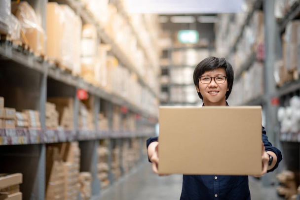Молодой азиатский счастливый человек с картонной коробкой между рядами полок на складе, в магазине или рабочий выбор и упаковка концепции
 - Фото, изображение