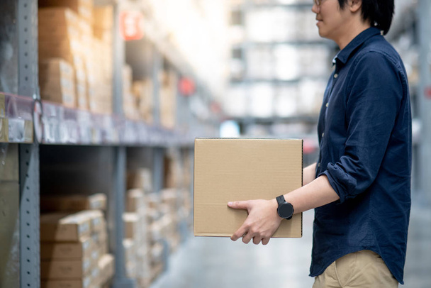 Young Asian man carry cardboard box between row of shelves in warehouse, shopping warehousing or working pick and packing concepts - Photo, Image
