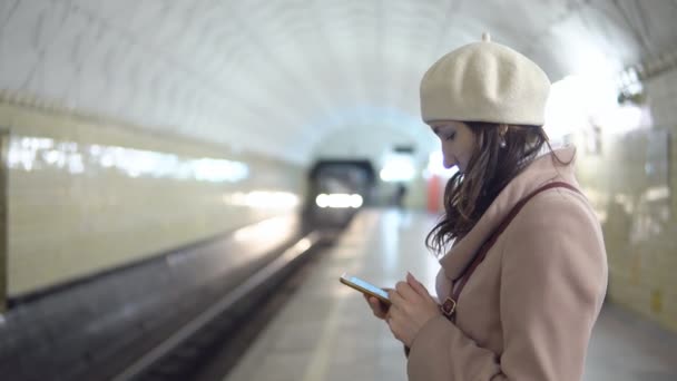 Młoda kobieta w płaszcz z telefonem w metropilitene czeka na pociąg - Materiał filmowy, wideo