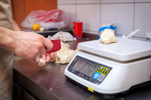 A close-up of a female baker  weighs on kitchen scales the dough for cooking buns on a knotted table with a rolling pin - Photo, Image
