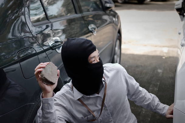 覆面をした強盗強盗の車を背景に小さな石を準備ができて保持目出し帽を身に着けています。保険犯罪概念. - 写真・画像