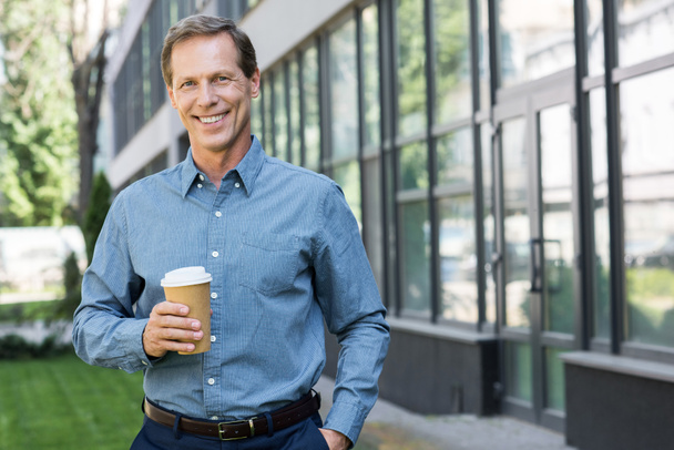 sonriente hombre de negocios posando con taza de café desechable cerca del edificio de oficinas
  - Foto, imagen