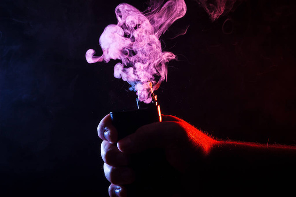 Close-up of a man holding a vape in his hand, which is smoking. Colorful smoke from cigarettes - Foto, immagini