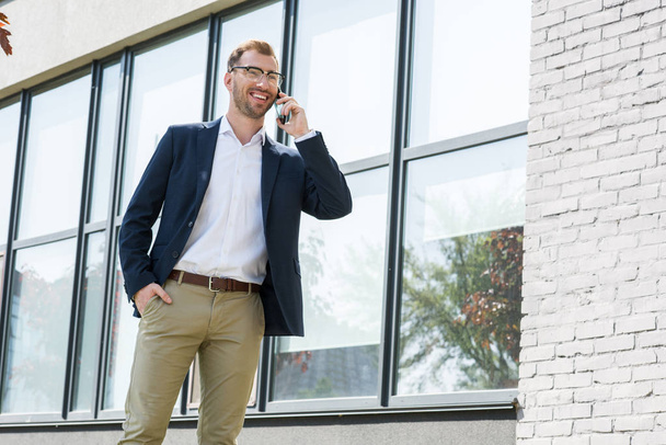 smiling businessman in formal wear talking on smartphone near office building  - Foto, imagen