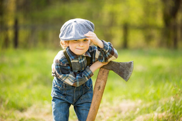 Small caucasian boy in hat posing with big axe. Portrait of small lumberjack - Zdjęcie, obraz