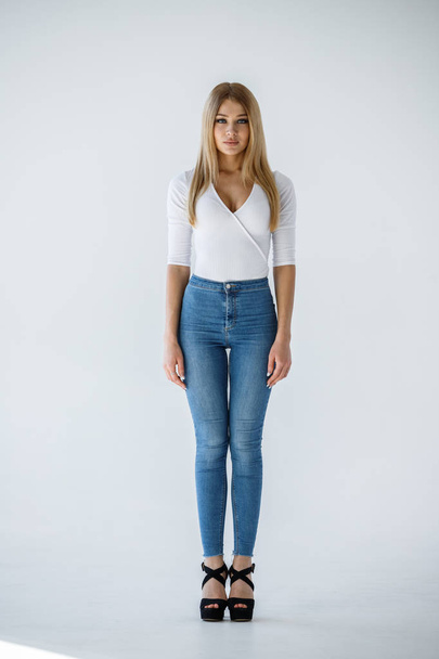 Studio shot of lovely blonde girl in white shirt and blue jeans standing on white background - Photo, Image