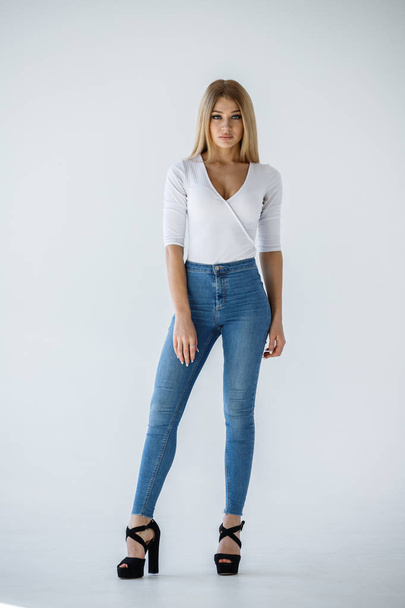 Studio shot of lovely blonde girl in white shirt and blue jeans standing on white background - Фото, изображение