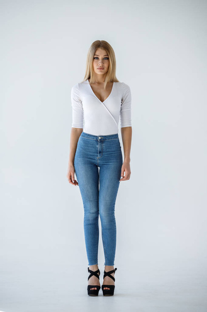 Studio shot of lovely blonde girl in white shirt and blue jeans standing on white background - Fotoğraf, Görsel