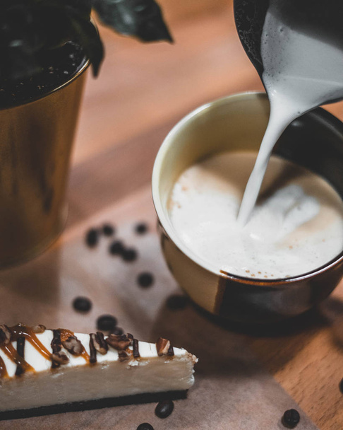 Closeup of milk pouring into coffee cup with coffee beans and cake on wooden board - Фото, зображення