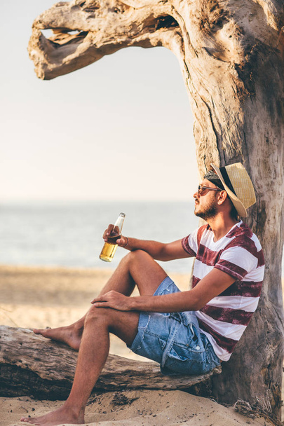 Young man sitting near dry tree and drinking beer on seacoast - Foto, Imagem