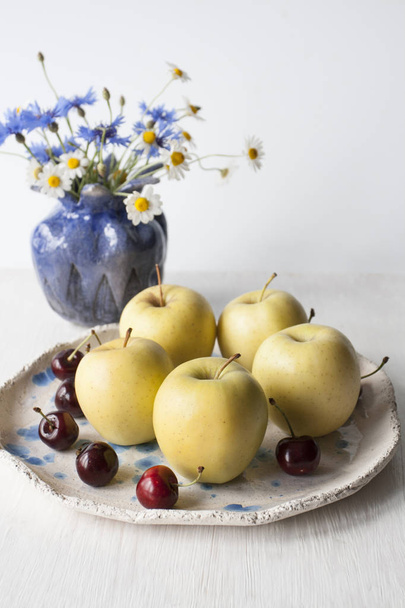 Des pommes jaunes et des cerises juteuses reposent sur une assiette d'argile. A proximité se trouve un vase à pigeons avec des fleurs de champ. Composition sur un fond en bois blanc
. - Photo, image