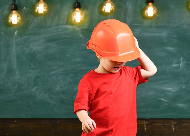 Мальчик играет как строитель или архитектор. Ребенок мечтает о будущей карьере в архитектуре или строительстве. Мальчик в оранжевой каске или шлеме, доска на заднем плане. Концепция детства
 - Фото, изображение