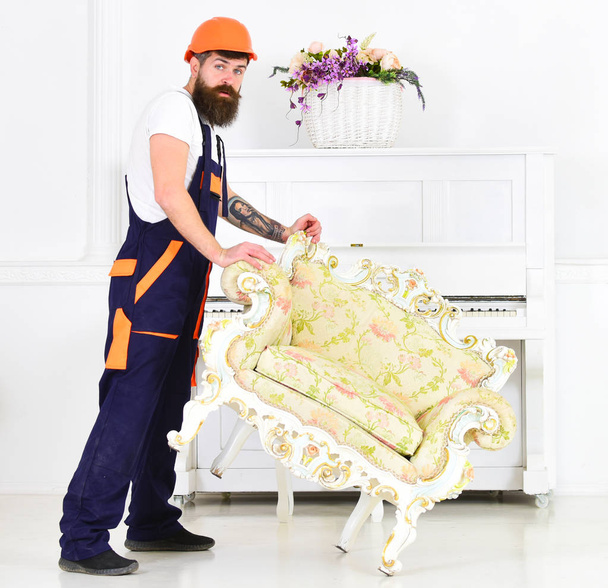 Relocating concept. Courier delivers furniture in case of move out, relocation. Man with beard, worker in overalls and helmet lifts up armchair, white background. Loader moves armchair for move out - Photo, image