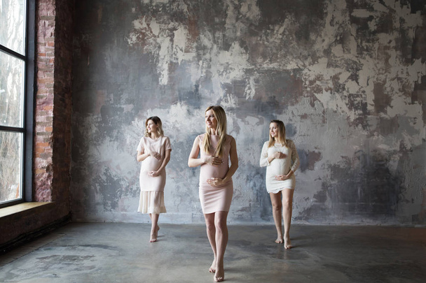 Три беременные женщины стоят с счастливыми улыбками и смотрят в большое окно. Одна беременная женщина в фокусе и две беременные женщины в дефокусе. Серая задняя стена
 - Фото, изображение