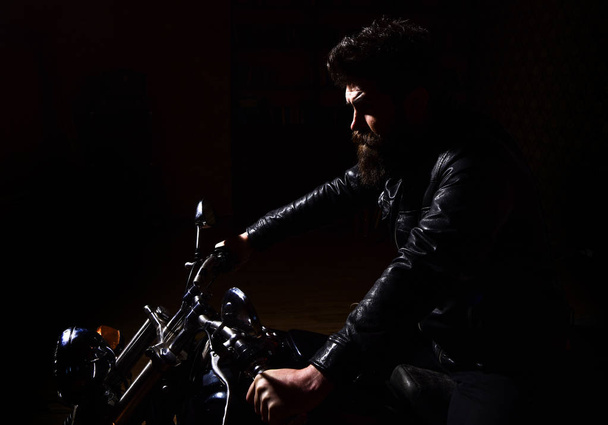 Masculinity concept. Man with beard, biker in leather jacket sitting on motor bike in darkness, black background. Macho, brutal biker in leather jacket riding motorcycle at night time, copy space - 写真・画像