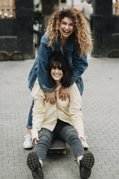 Full length portrait of two girlfriends having fun while one girl is sitting on a skateboard and another one is pushing her in the back. - Фото, изображение