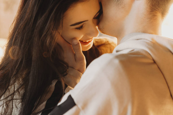 Bonito close up retrato de um jovem casal caucasiano abraçando contra a luz, enquanto o menino está beijando sua bochecha namorada que está sorrindo
. - Foto, Imagem
