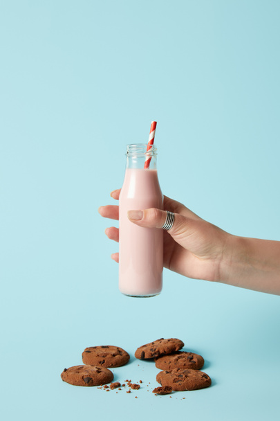 cropped shot of woman holding strawberry milkshake in bottle with drinking straw and chocolate cookies on blue background  - Photo, Image