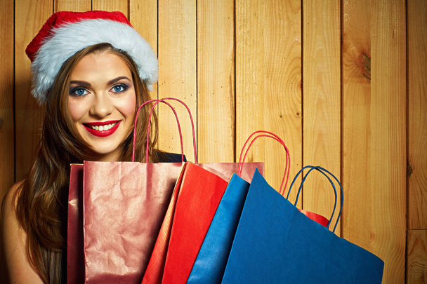 mujer sonriente con sombrero de Santa sosteniendo bolsas de compras sobre fondo de madera
  - Foto, imagen