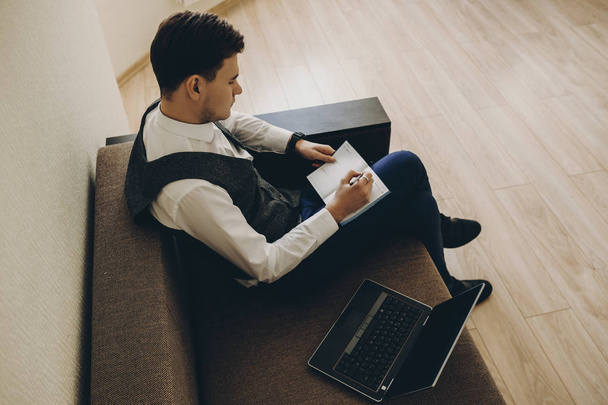 Верхний вид молодого кавказского менеджера в очках, сидящих на диване и работающего на ноутбуке, отмечая в ежедневном журнале
. - Фото, изображение