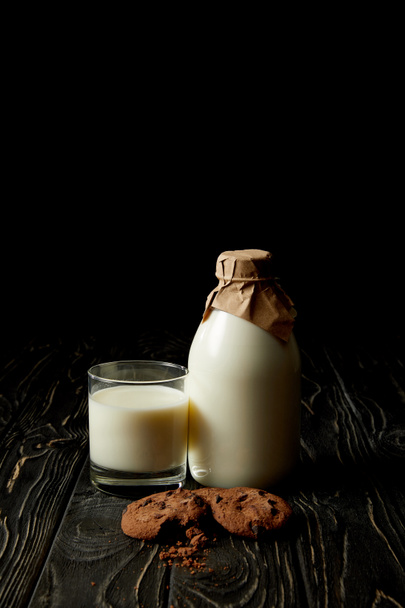 φρέσκο γάλα στο ποτήρι, μπουκάλι γάλα που είναι τυλιγμένο με χαρτί και μπισκότα σοκολάτας σε μαύρο φόντο  - Φωτογραφία, εικόνα