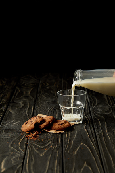 περικομμένη εικόνα πρόσωπο ρίχνει το γάλα σε ένα ποτήρι από το μπουκάλι και μπισκότα σοκολάτας σε μαύρη ξύλινη επιφάνεια - Φωτογραφία, εικόνα