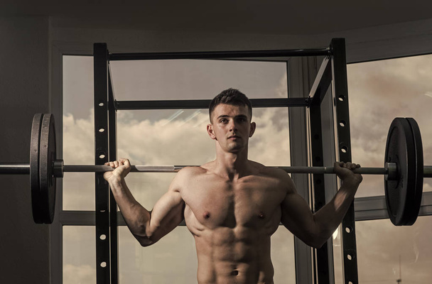 Sport and gym concept. Man with torso, muscular macho does exercise with barbell, window on background. Sportsman, athlete looks attractive. Man with nude torso in gym enjoy his sporty lifestyle - Fotó, kép