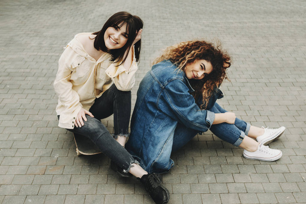 Portrait of two sisters dressed in jeans blue and yellow jackets sitting on the pavement and looking at camera. - Fotó, kép