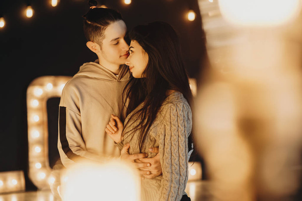 Jeune et beau couple caucasien embrassant et regardant vers l'autre avec amour à travers des lumières chaudes dans une chambre noire décorée de lumières chaudes
. - Photo, image