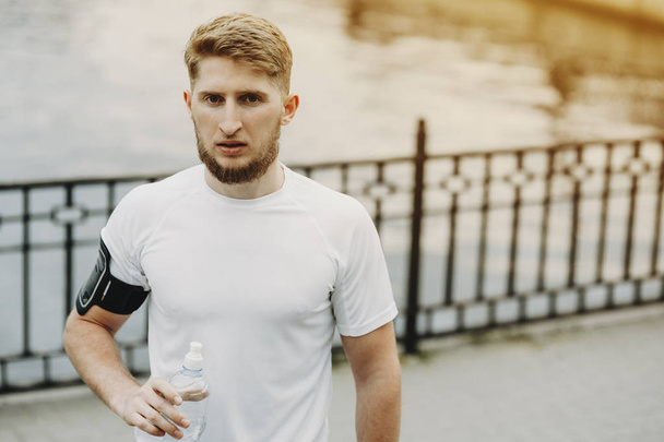 Porträt eines gesunden Mannes, der nach dem Laufen im Freien mit einer Flasche Wasser in die Kamera blickt. - Foto, Bild